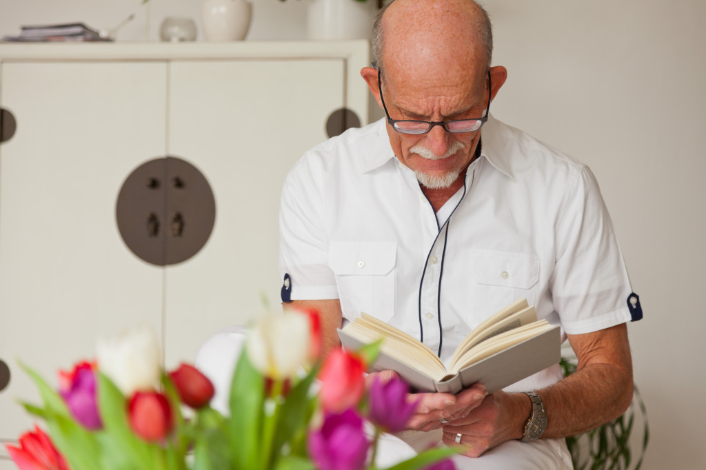 Senior man with glasses reading book in living room.