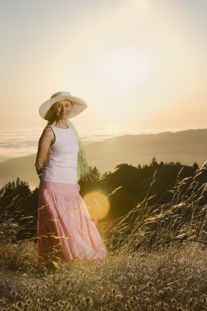 Senior woman in cute floppy hat wearing loose fitting top and long flowing skirt while standing in a grassy field at sundown. 