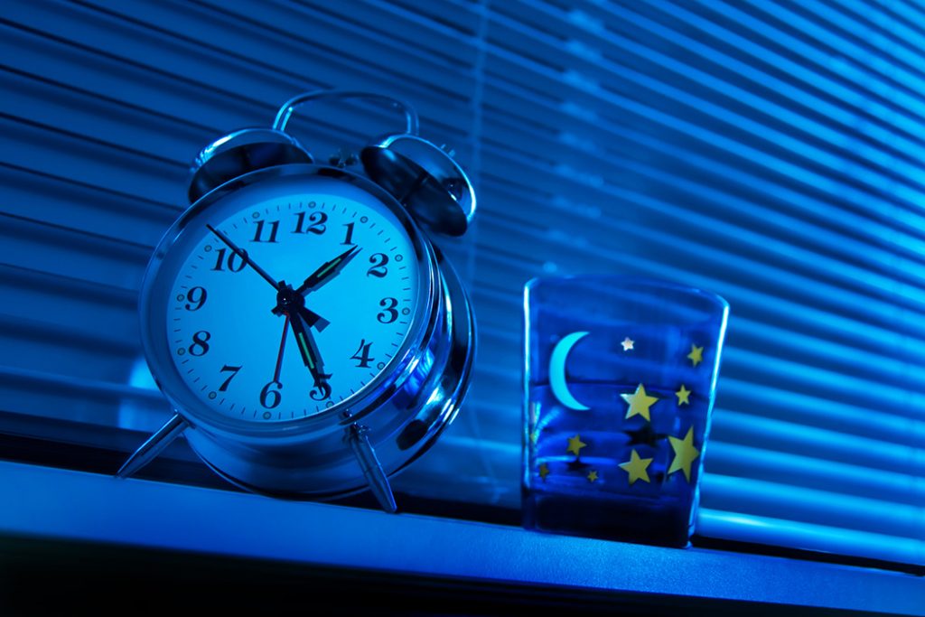 Alarm clock with water glass on night stand in dark blue room.