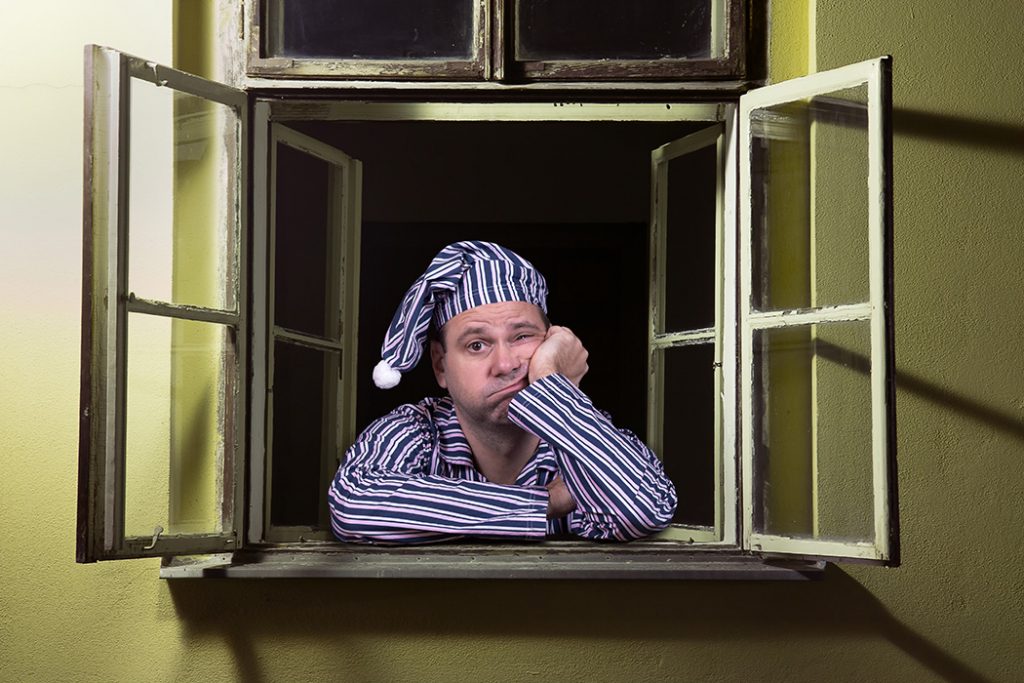 Man in night shirt and cap wide awake and looking sadly out bedroom window at night.