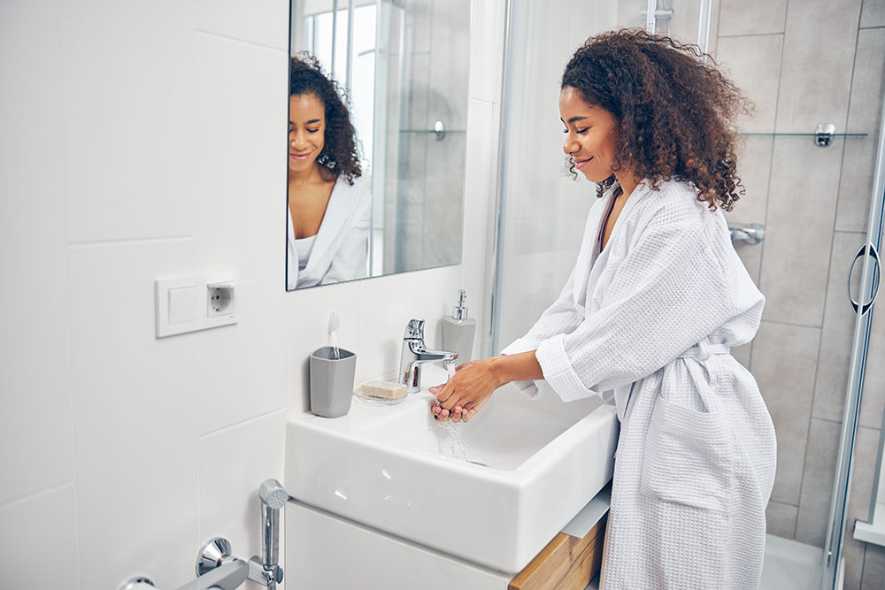 Young woman at home in white bathrobe washing hands in sink. in the bathroom