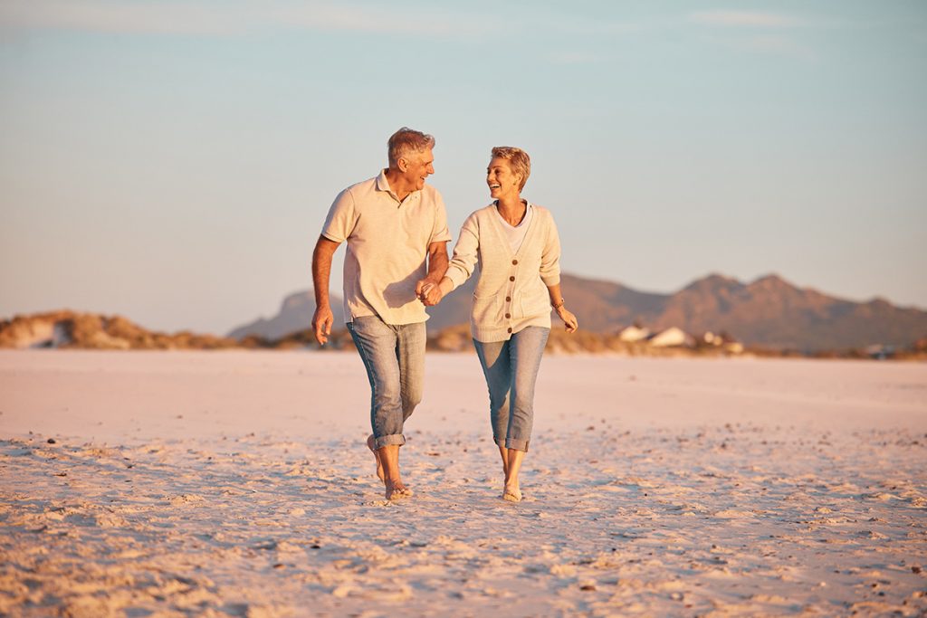 Lovely senior couple smiling and holding hands as they walk on the beach enjoying a sunny day.