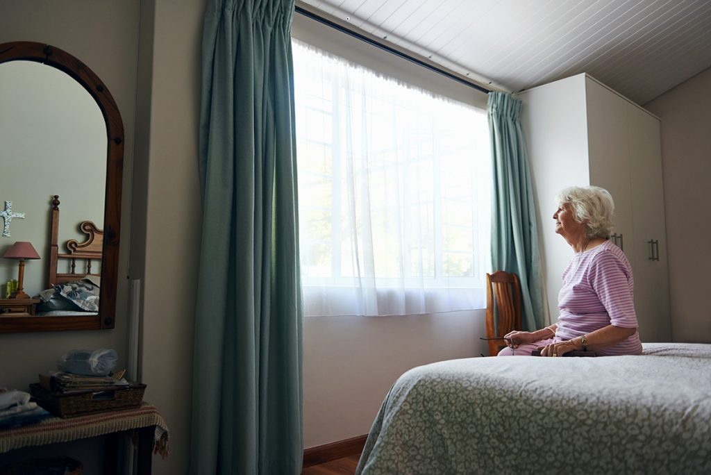 Senior woman sitting on bed in bedroom looking out the window.