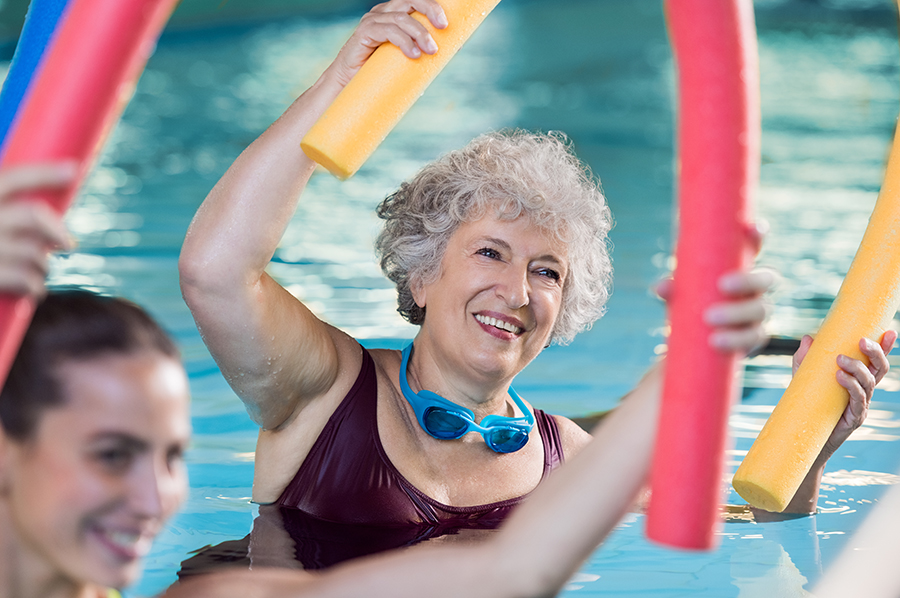 Smiling senior woman doing aqua fitness with swim noodles. Happy mature healthy woman taking fitness classes in aqua aerobics. Healthy old woman holding swim noodles in hand doing aqua gym with young trainer.