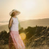 Senior woman in cute floppy hat wearing loose fitting top and long flowing skirt while standing in a grassy field at sundown.