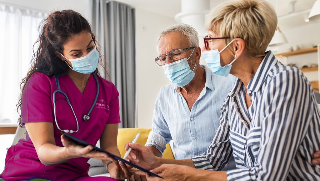 Female nurse talking to seniors patients with mask while being at home.