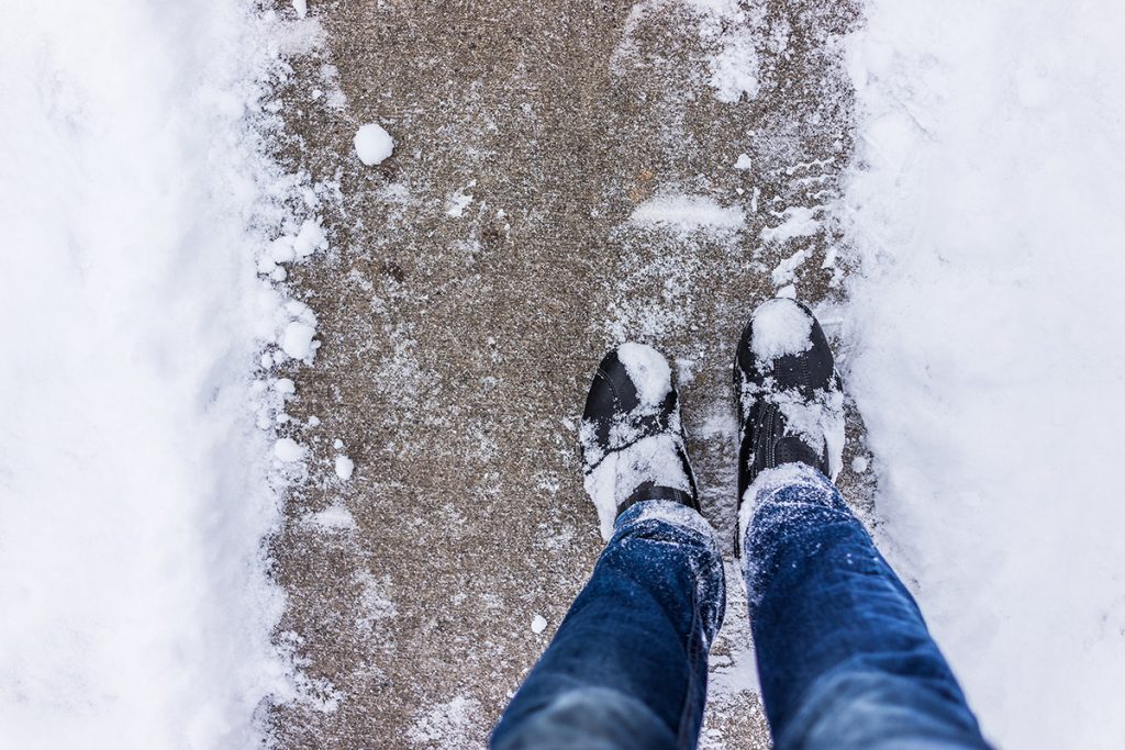 Senior wearing snowy boots and jeans looking down at shoveled walkway.